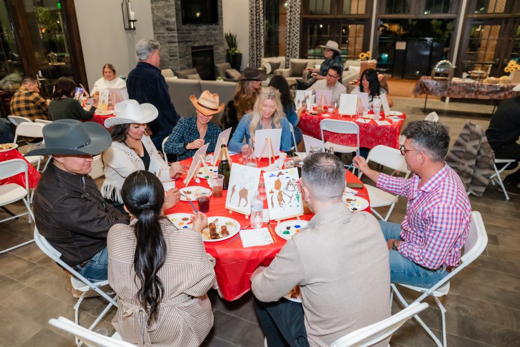 A group of people wearing cowboy hats and casual clothes sit around tables with red tablecloths, painting canvases. Paint palettes, brushes, and drinks are on the tables in a brightly lit room with large windows.