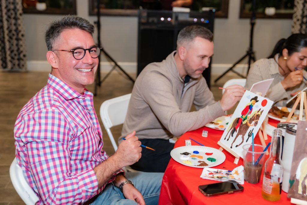 A smiling man in a pink plaid shirt sits at a table, painting on a canvas, while two others beside him focus on their artwork. The table holds paint supplies, drinks, and a smartphone.