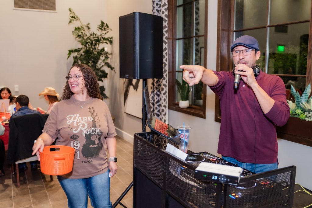 A man in a cap speaks into a microphone and points while standing behind DJ equipment. A woman nearby holds an orange raffle basket and smiles. People sit at tables in the background of the indoor event.