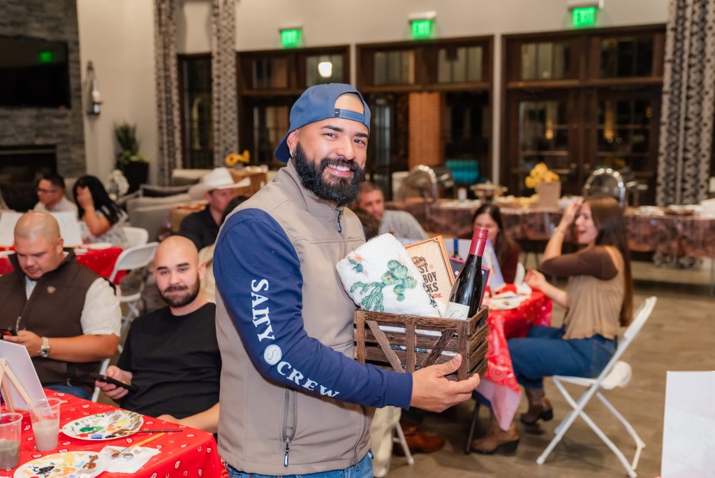 A smiling man in a cap and vest holds a gift basket with wine and towels in a room with people seated at tables covered with red tablecloths, enjoying a festive indoor gathering.