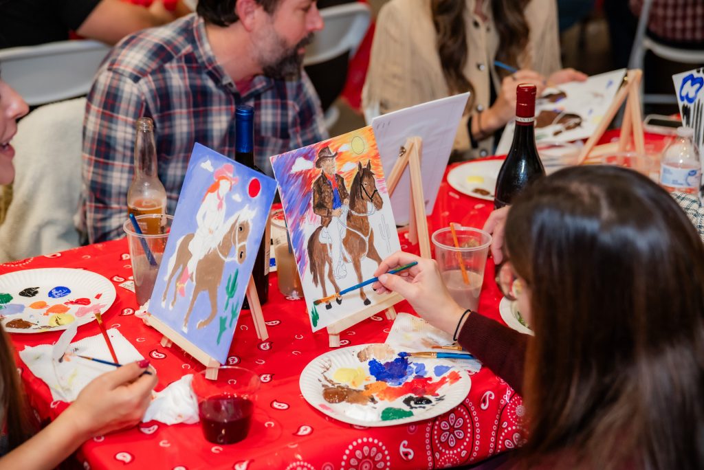A group of people sit at a table covered with a red bandana cloth, painting cowboy-themed artwork on canvases. Paint supplies, drinks, and bottles are scattered on the table.