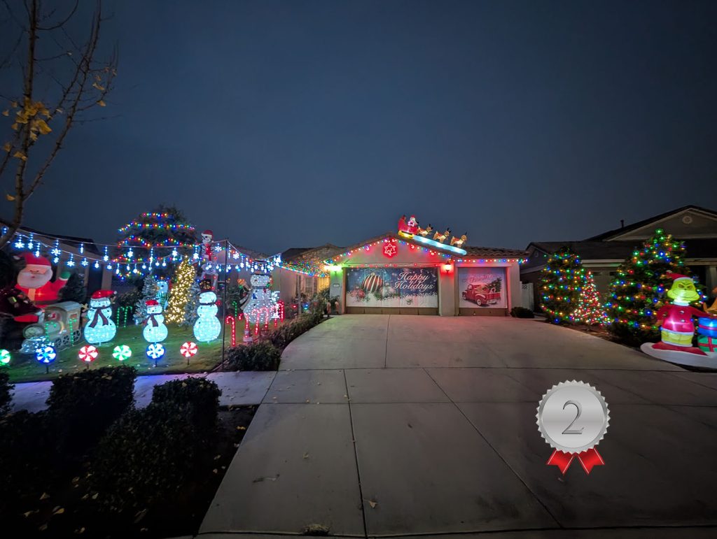 A house decorated with colorful Christmas lights and inflatables, including Santa, snowmen, and a tree. The roof features a Santa and reindeer display. A silver ribbon with a "2" badge appears in the lower right corner.