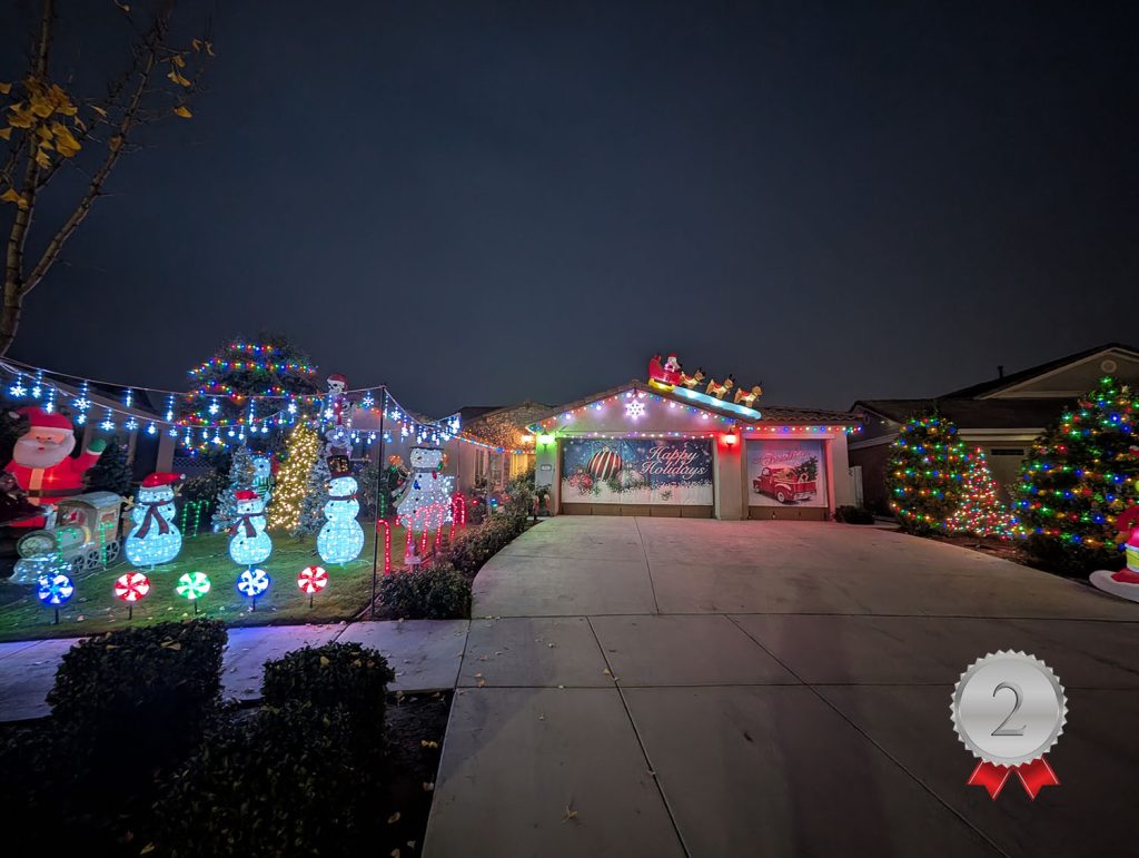 A house decorated with colorful Christmas lights, inflatable snowmen, Santa figures, and a sleigh with reindeer on the roof. The yard features glowing decorations and two brightly lit trees. A "2" ribbon emblem is at the bottom right.