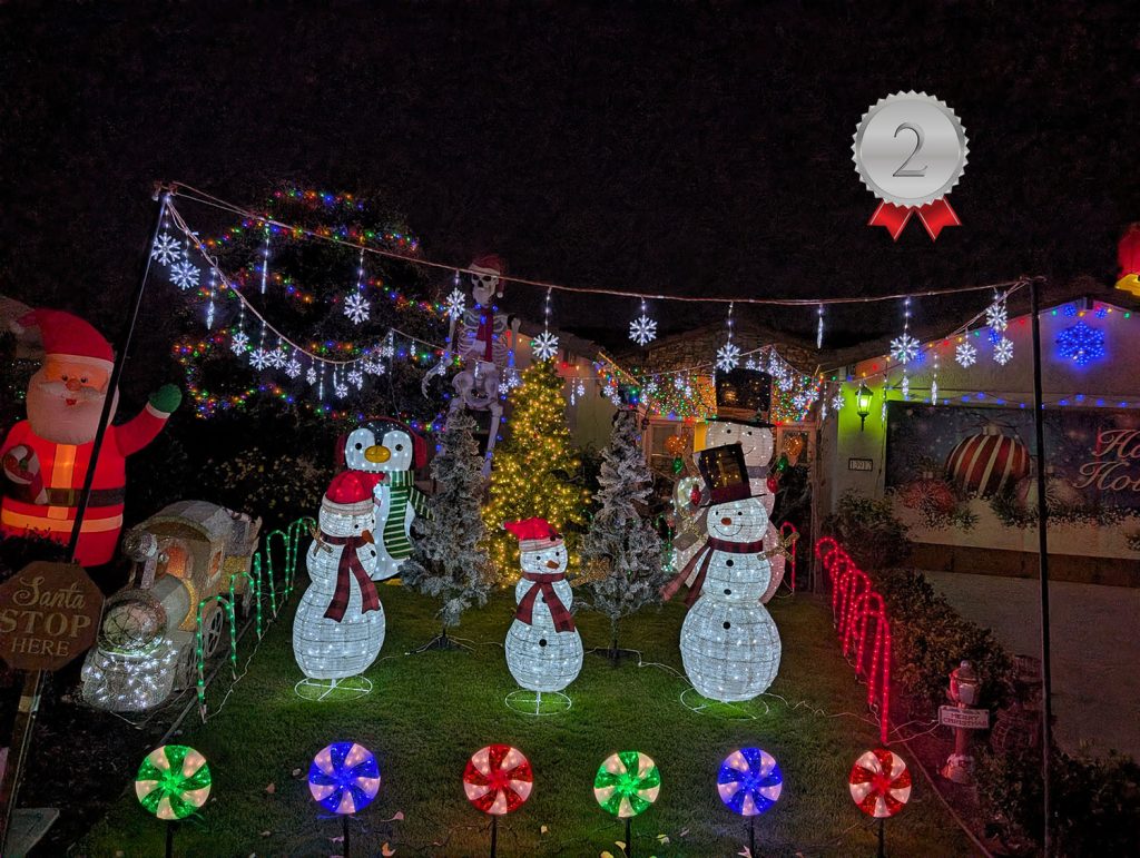 A house decorated for Christmas with glowing snowmen, candy cane lights, string lights, and an inflatable Santa. A “2nd place” ribbon is displayed in the top right corner.