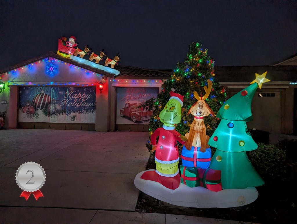 A house decorated with colorful Christmas lights and inflatables, including the Grinch, Max, and a Christmas tree. The garage door has festive murals, and Santa with reindeer is on the roof. A "2" silver ribbon is in the corner.