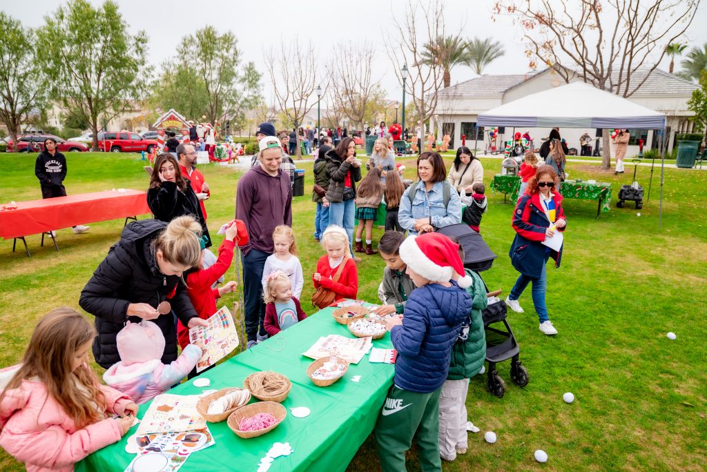 Children and adults participate in a festive outdoor event with activity tables, crafts, and holiday decorations on green grass, surrounded by trees and a white tent. Some children wear Santa hats and winter clothes.