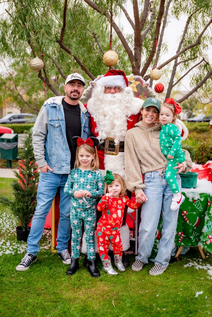A family with two adults and three young children in festive pajamas poses outdoors with Santa Claus. Christmas decorations and trees are in the background, creating a cheerful holiday scene.