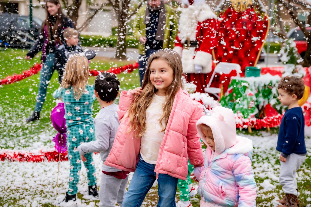 Children play outside in artificial snow, wearing colorful winter jackets. A girl in a pink coat smiles at the center, with festive decorations and adults in the background, including someone dressed as Santa Claus.