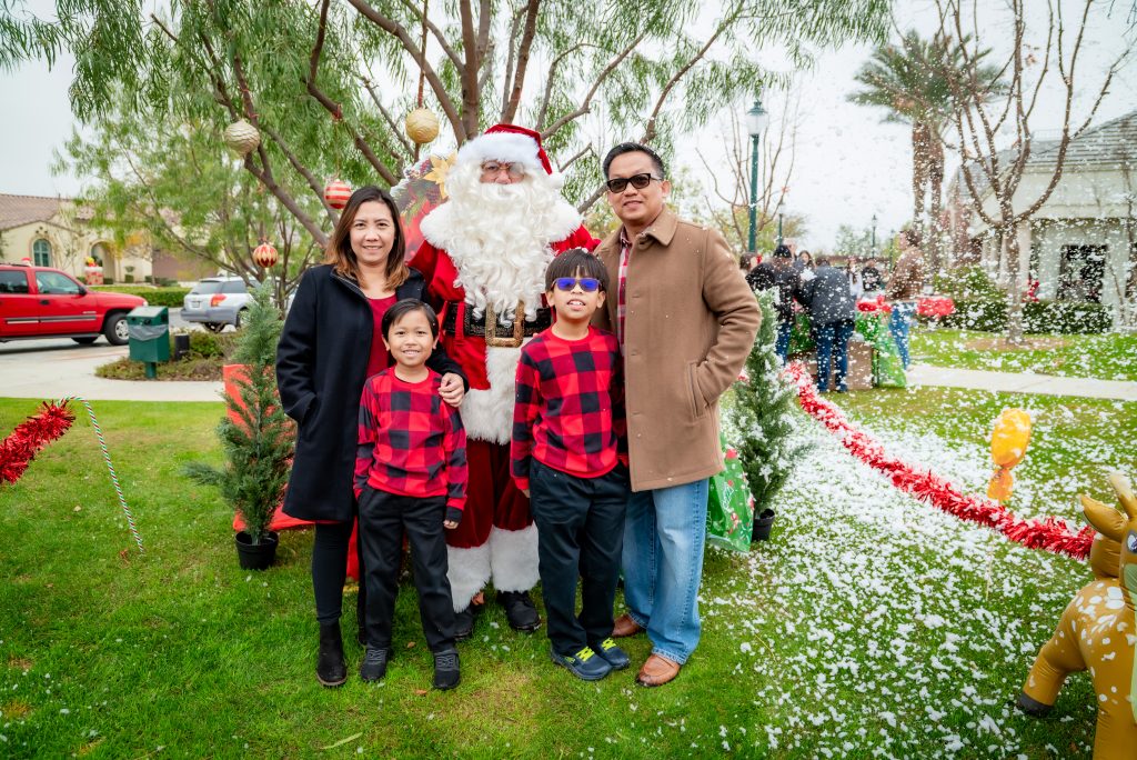 A family of four poses outdoors with Santa Claus, surrounded by artificial snow, small decorated trees, and festive holiday decorations. The children wear matching red plaid shirts, and everyone is smiling.