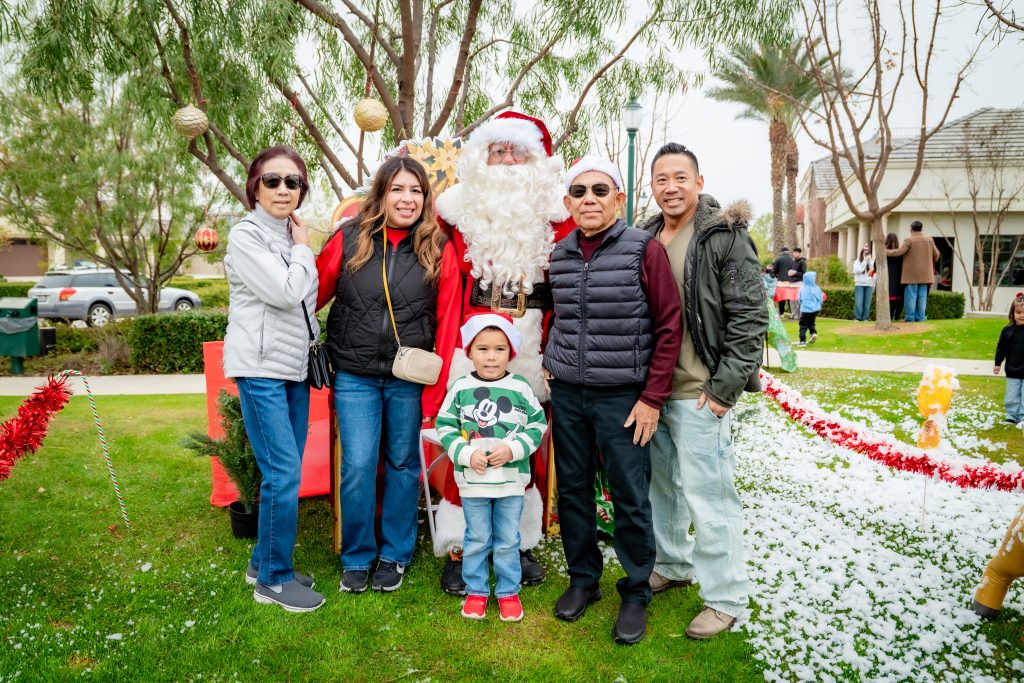 Six people and a child pose outdoors with Santa Claus in a festive holiday setting, surrounded by greenery, decorations, and artificial snow. All are smiling and dressed in casual winter clothing.