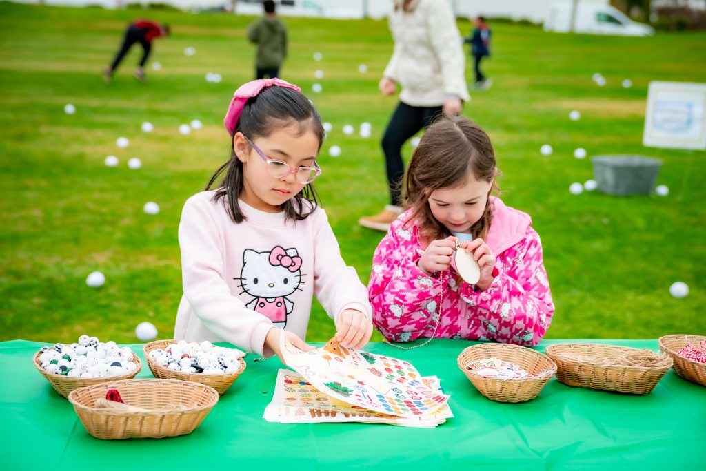 Two young girls sit at a green table outdoors, surrounded by baskets of craft supplies. One girl looks at stickers while the other holds a decorated item. People and white balls are scattered on the grass in the background.