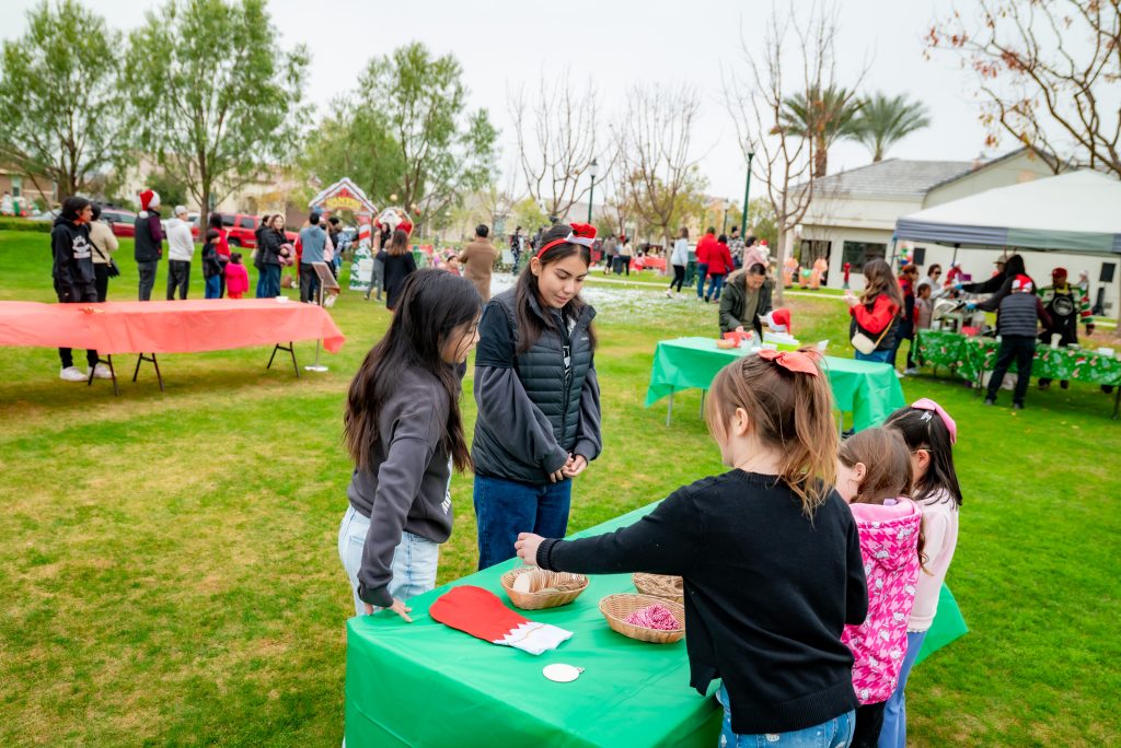 Children gather around a green-covered table at an outdoor holiday event, participating in crafts. Other groups and decorated booths are visible in the background on the grassy park.