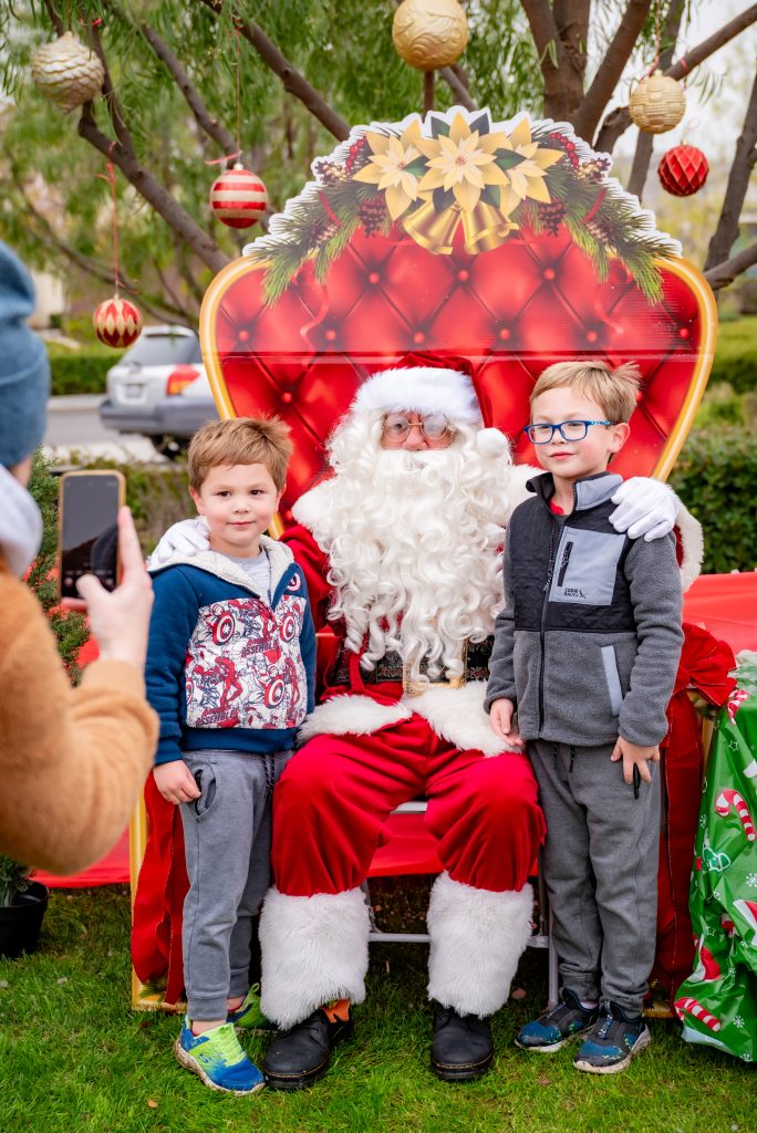 Two young boys stand next to Santa Claus, who is seated on a festive red chair outdoors, as someone takes their photo. Christmas ornaments hang from the tree behind them, creating a holiday atmosphere.