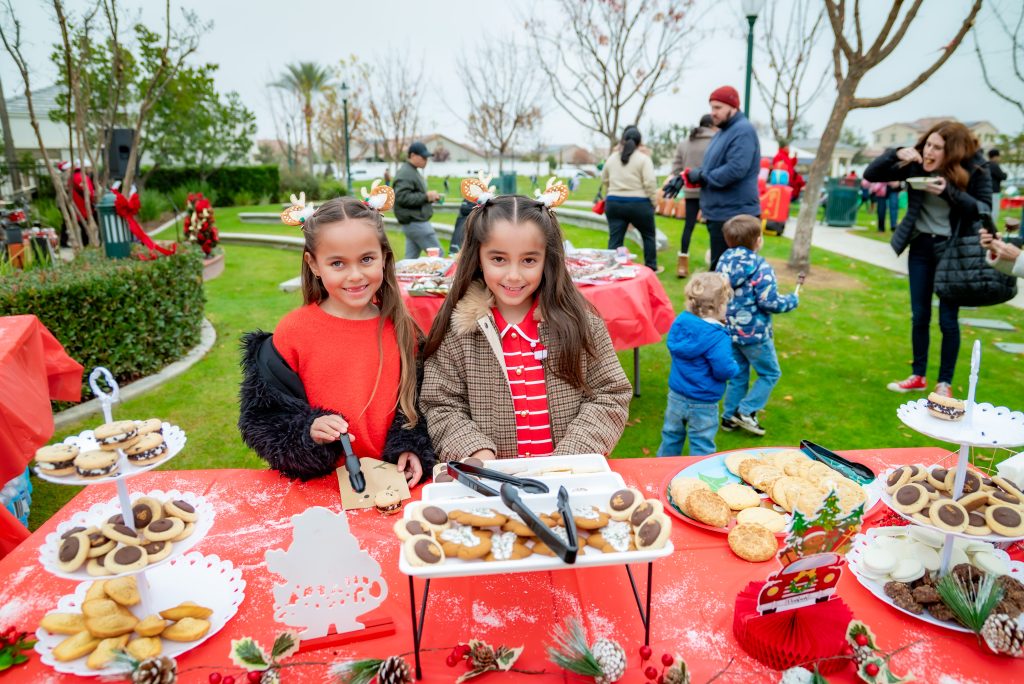 Two smiling girls stand at a festive outdoor table filled with cookies and treats. People and children are in the background, and the scene is decorated for the holidays with red tablecloths and pine cone centerpieces.