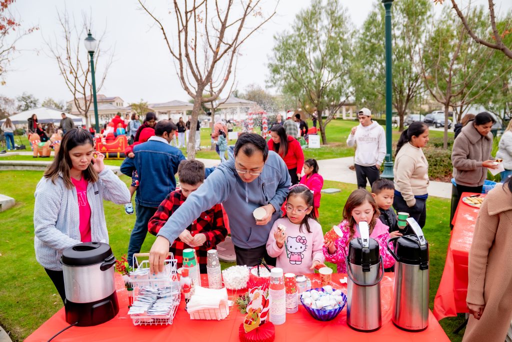 A group of people gathers at an outdoor event in a park, serving themselves snacks and drinks from a red-covered table. Children and adults are present, and trees and houses are visible in the background.
