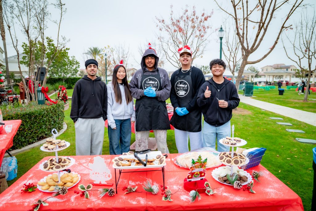 Five young people in festive hats and aprons stand behind a table with holiday cookies at an outdoor event in a park, smiling at the camera. Red tablecloths and holiday decorations are visible.