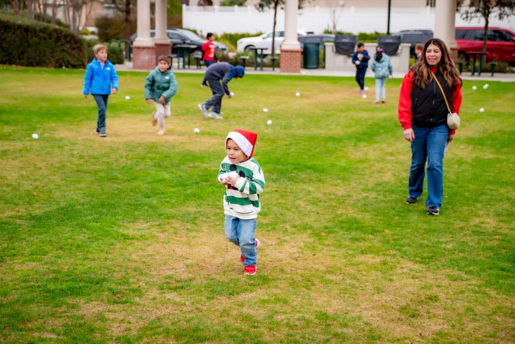 A young child wearing a Santa hat runs on grass holding a snowball, followed by a smiling woman and other children playing in the background at a park.