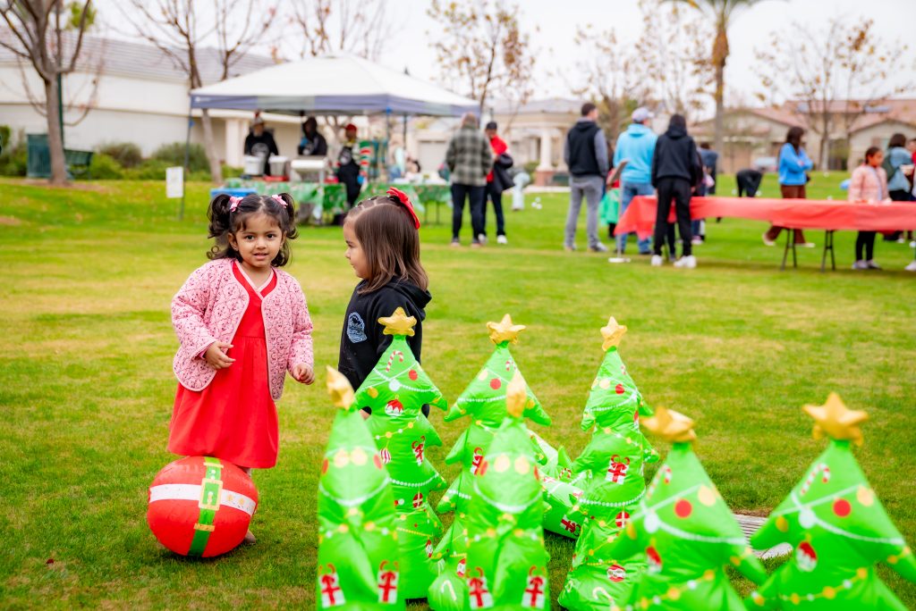 Two young girls stand on the grass near inflatable Christmas trees and a large holiday-themed ball, with people gathered at tables and under a canopy in the background at an outdoor event.