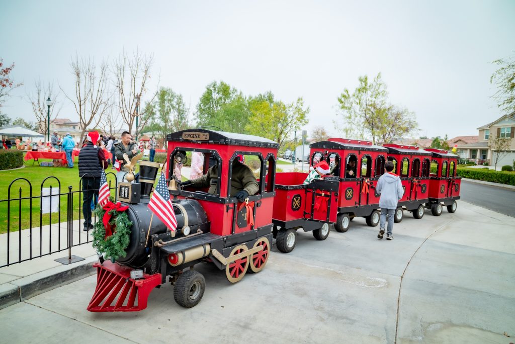 A small red and black trackless train decorated with a wreath and American flags gives rides to people at a park. Several passengers and a driver in Santa hats are visible, with trees and houses in the background.
