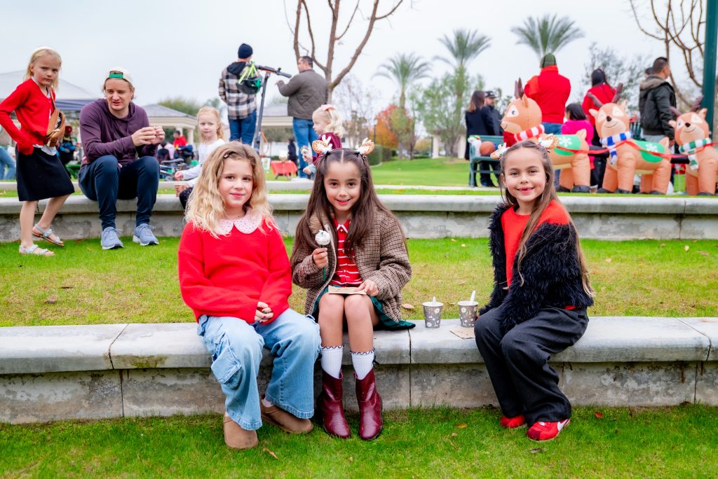 Three young girls wearing festive outfits sit on a concrete bench outdoors, smiling. Behind them, people and holiday decorations, including inflatable reindeer, are visible on a grassy area.