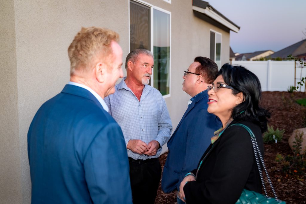 Four adults in business attire are standing outside a house, engaged in conversation near a beige wall and windows, with landscaping in the background.