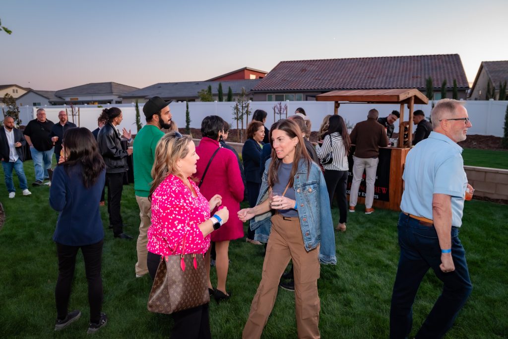 A group of people socialize in a backyard setting at sunset. Two women in the foreground talk, one in a pink blouse and the other in a denim jacket. Others mingle near a wooden drink stand on the grass.