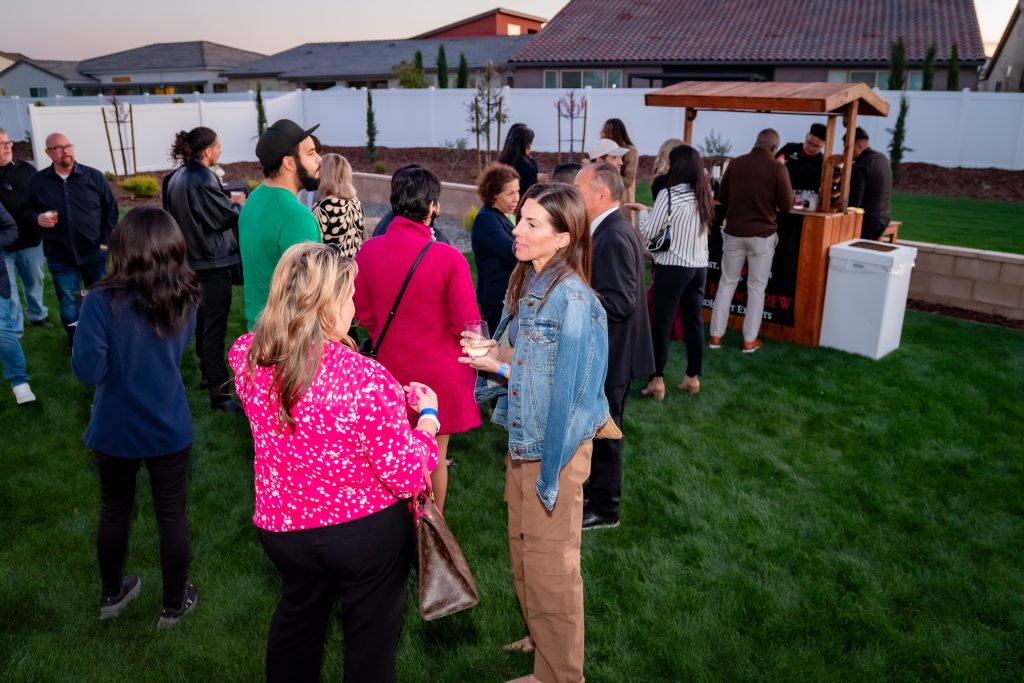 A group of people socialize at an outdoor gathering on a lawn, with some standing near a wooden bar. Houses and a white fence are visible in the background. Most people are chatting and holding drinks.