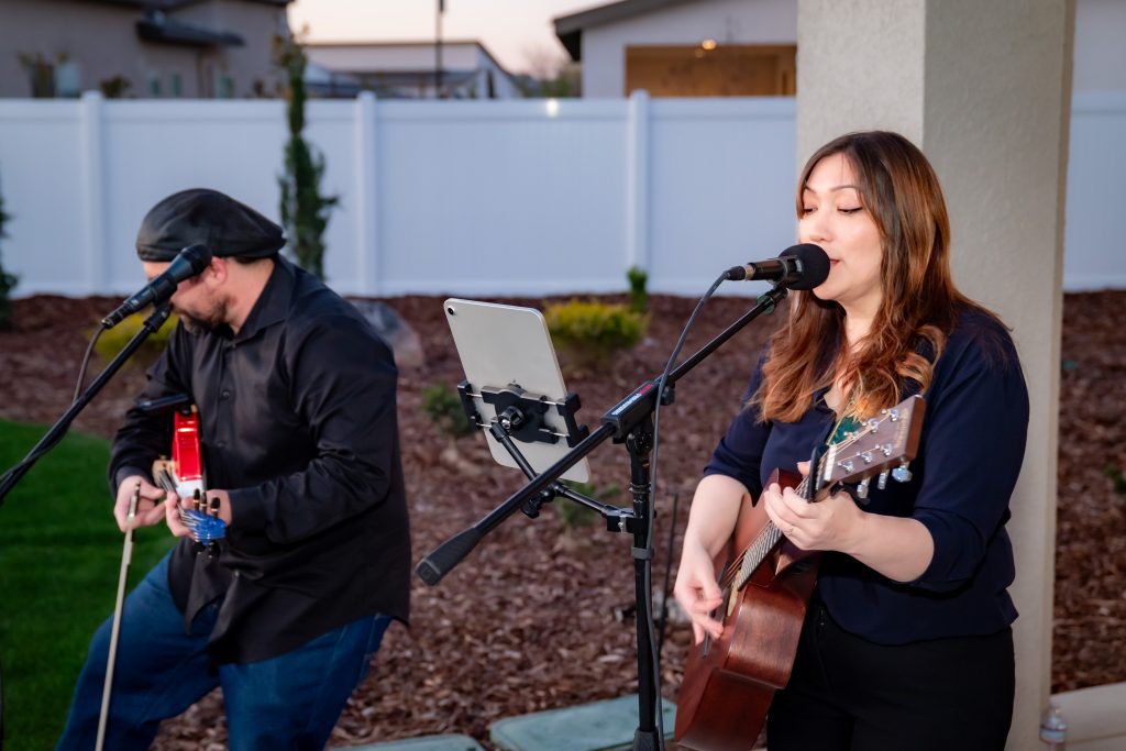 A woman sings and plays acoustic guitar into a microphone while a man plays electric guitar beside her outdoors, near a white fence and houses. An iPad is mounted on a stand in front of them.