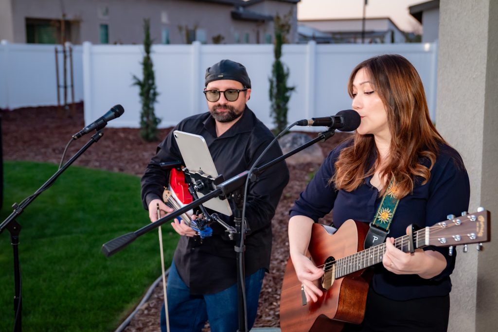 A woman sings and plays an acoustic guitar while a man beside her plays an electric guitar and reads from a tablet, performing outdoors in a backyard with a white fence and grass.