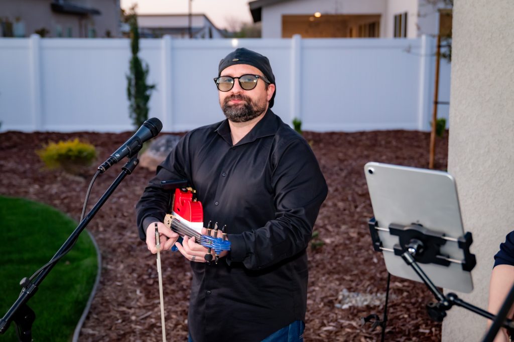 A man in a black shirt and glasses stands outdoors holding a violin near a microphone, with an iPad on a stand beside him and houses and a white fence in the background.
