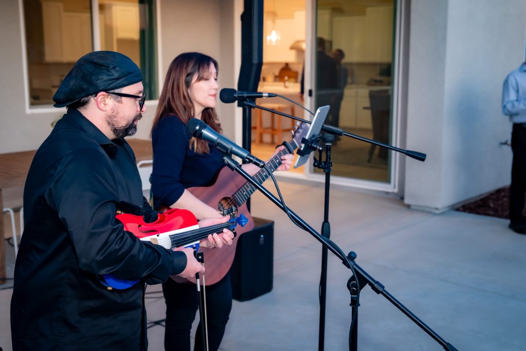Two musicians perform outdoors; a man plays an electric violin while a woman sings and plays guitar. Both are dressed in black, standing near microphones, with a house and patio visible in the background.
