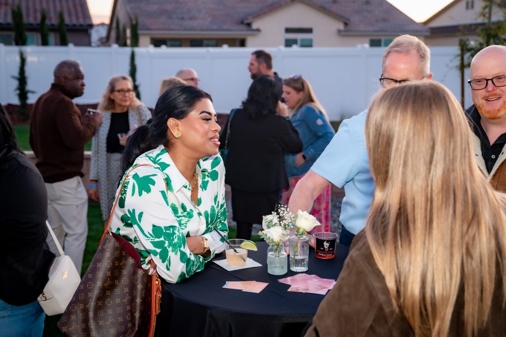 A woman in a white and green floral blouse talks and smiles with others around a small table at an outdoor social gathering. People are mingling in the background, and the event appears casual and friendly.
