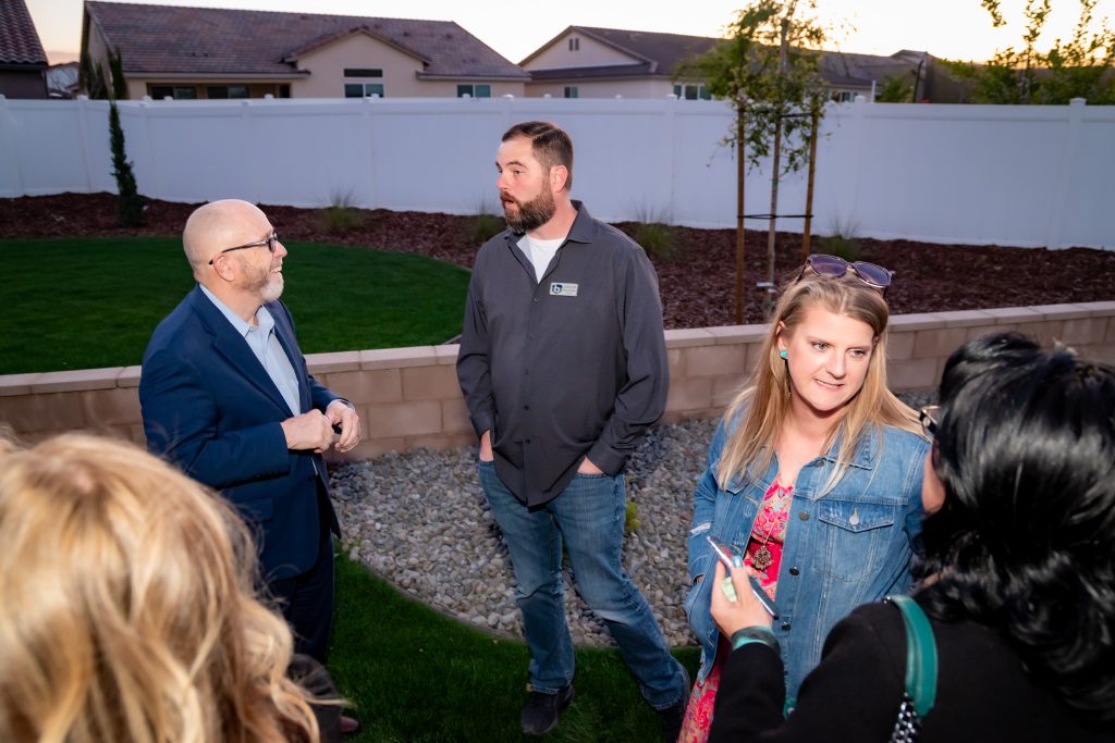 Four adults stand and talk in a suburban backyard with green grass, a rock border, and a white fence. Two men converse to the left, while two women chat to the right. Houses and trees are visible in the background.