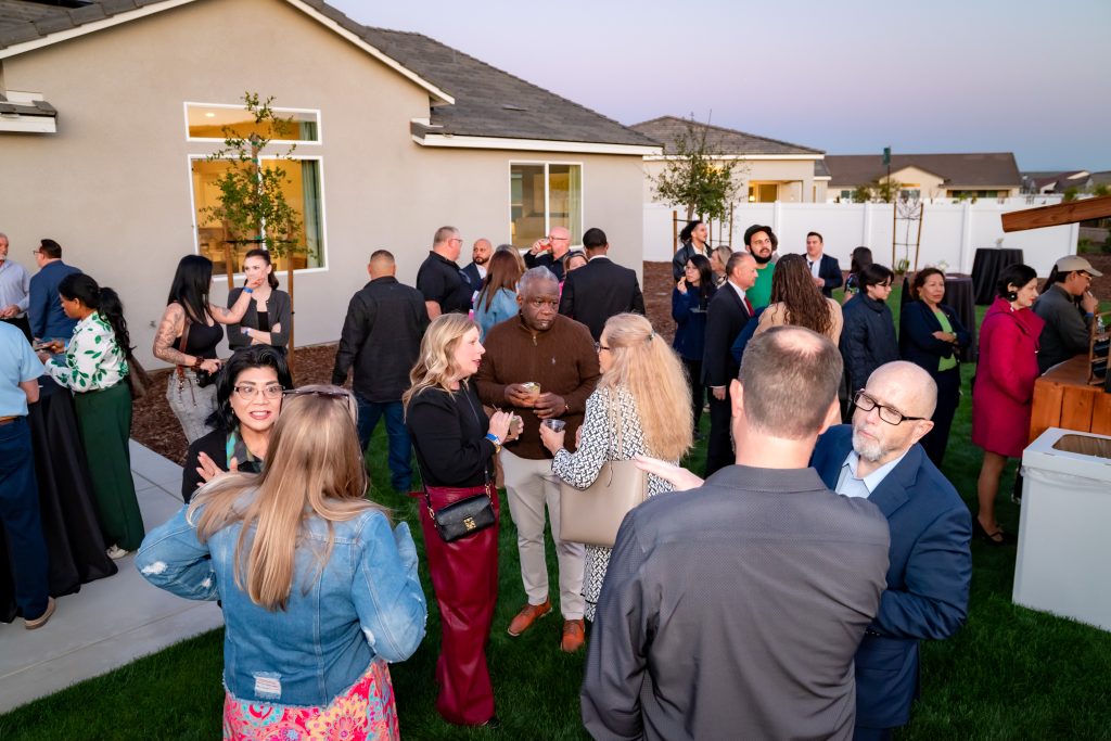 A group of people socialize outdoors at a backyard gathering, chatting in small groups near a modern house at dusk. Most are dressed in business or semi-formal attire. The atmosphere appears lively and friendly.