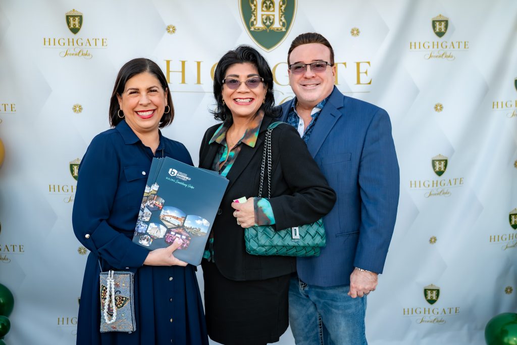 Three smiling adults pose together at an event, standing in front of a Highgate-branded backdrop. One woman holds a brochure, and all are dressed in smart-casual attire.