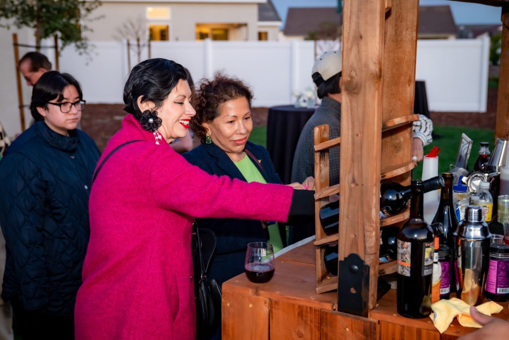Two women, one in a bright pink coat and the other in a teal coat, select a bottle of wine from a wooden outdoor bar. A wine glass and various bottles are on the counter. Other people are in the background.