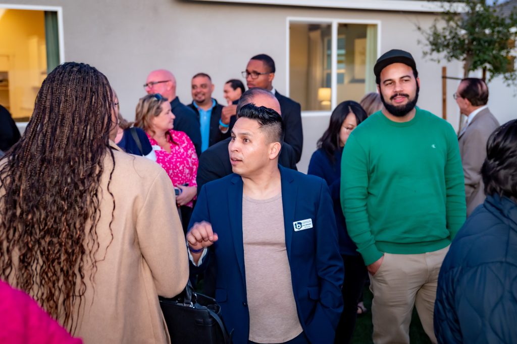A group of people gather outdoors at an event. One man in a blue blazer with a nametag is talking to a woman with braided hair. Others are standing and chatting in the background, some smiling.