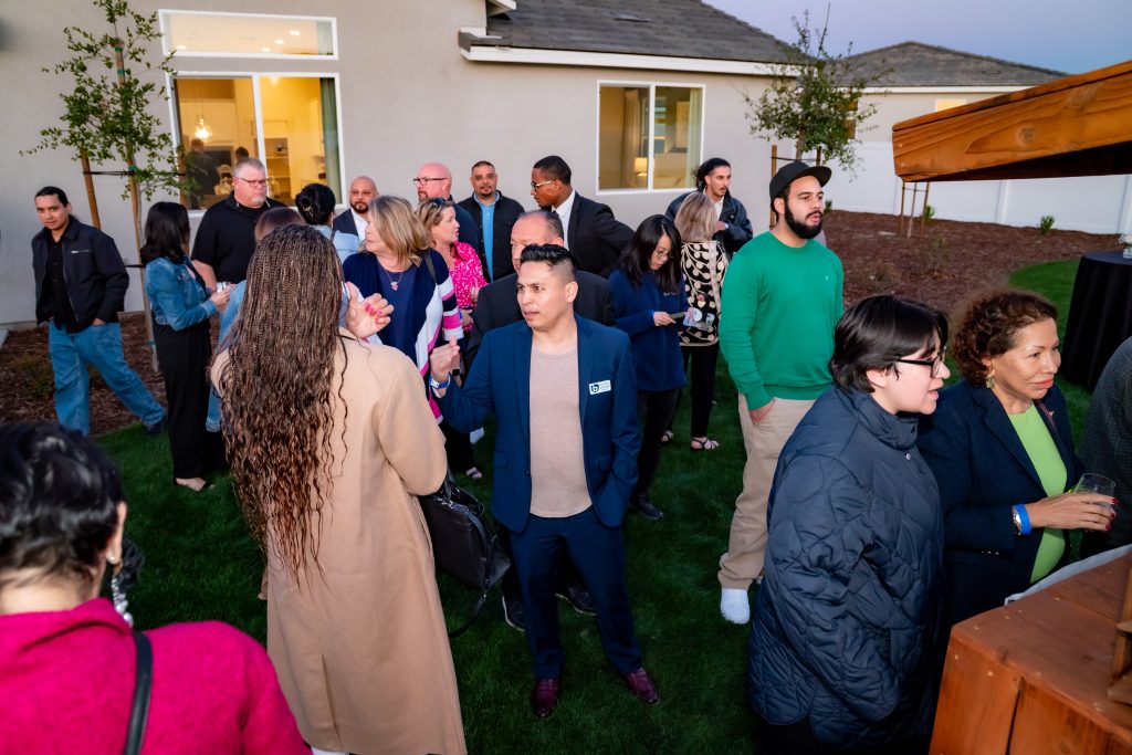 A group of people gather outdoors on a lawn for an event near a modern house at dusk, talking and socializing. Most are dressed in business or casual attire.