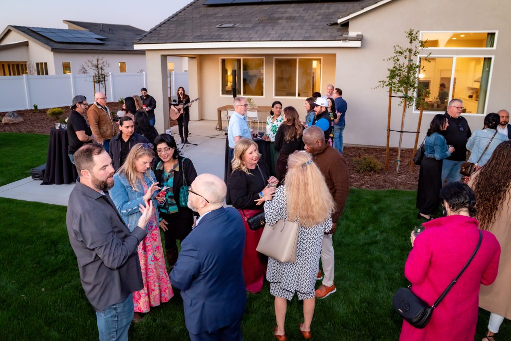 A group of people gather and socialize in the backyard of a house during an outdoor event, dressed in semi-formal attire, standing on the grass and near a patio as evening approaches.