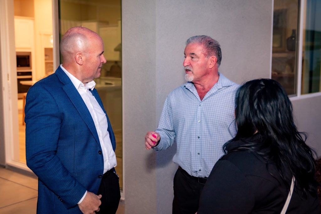 Three adults stand and talk outside near a building at night. Two men, one in a blue jacket and one in a checked shirt, face each other, while a woman with long dark hair stands nearby, listening.
