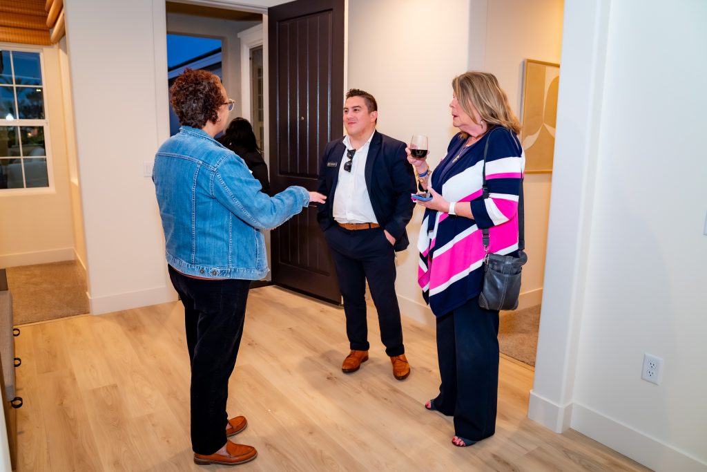 Three people are standing and talking in a bright hallway. One woman wears a denim jacket, another wears navy and a colorful striped shawl, and a man in a suit stands between them. The woman on the right holds a glass of wine.