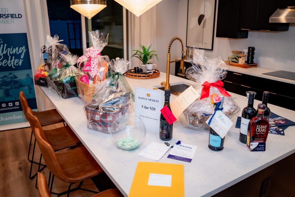 A kitchen island displays several gift baskets wrapped in cellophane, bottles of wine and whiskey, a glass bowl with raffle tickets, and envelopes, possibly prepared for a raffle or fundraising event.