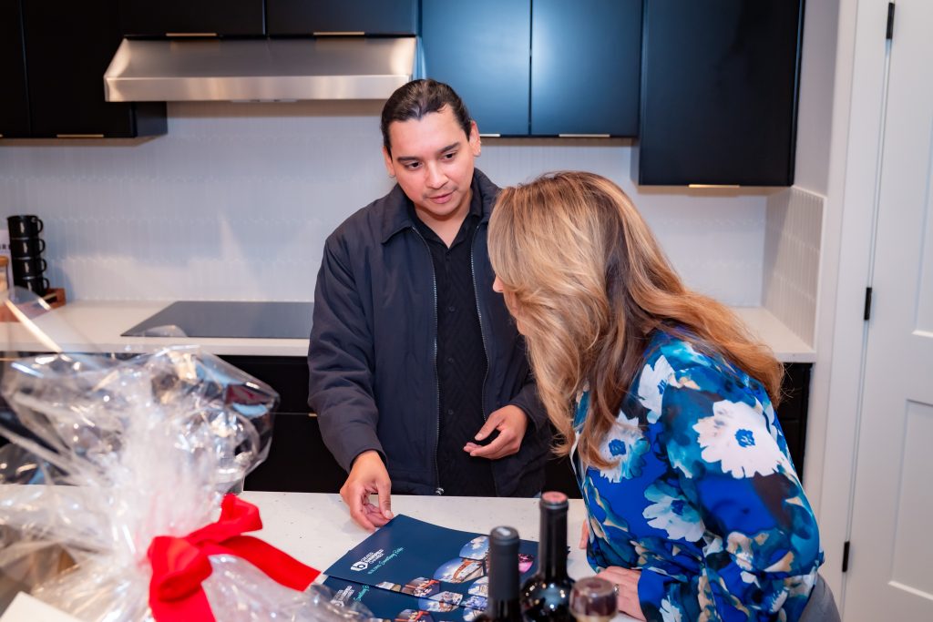 A man and woman stand in a modern kitchen, looking at a brochure on the counter. The man is pointing at the brochure while the woman leans in. Gift baskets and bottles of wine are visible in the foreground.