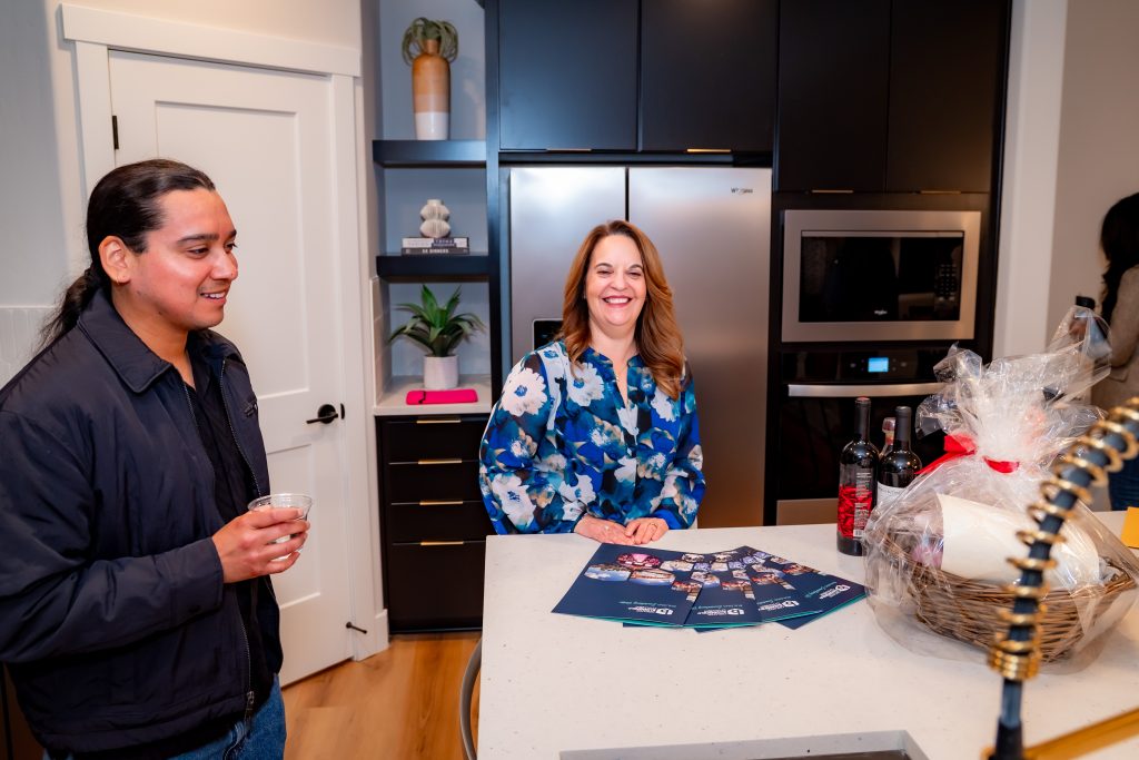 Two people stand and smile in a modern kitchen near a counter with brochures, wine bottles, and a gift basket. One person holds a drink, while the other stands behind the counter. Shelves and appliances are in the background.