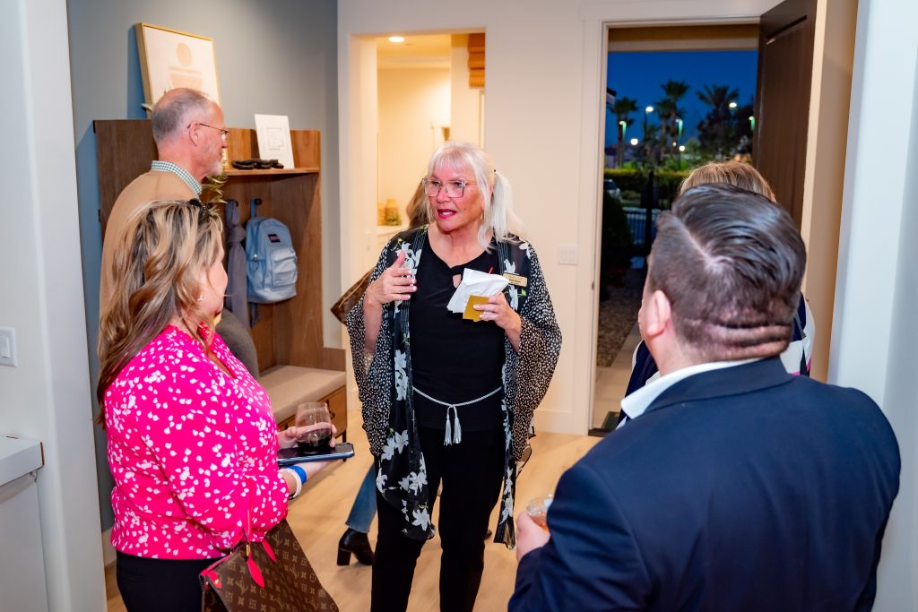 A group of adults stand chatting in a modern, well-lit room. One woman in the center gestures while speaking. Others listen, holding drinks, and palm trees are visible through the open door to the outside.