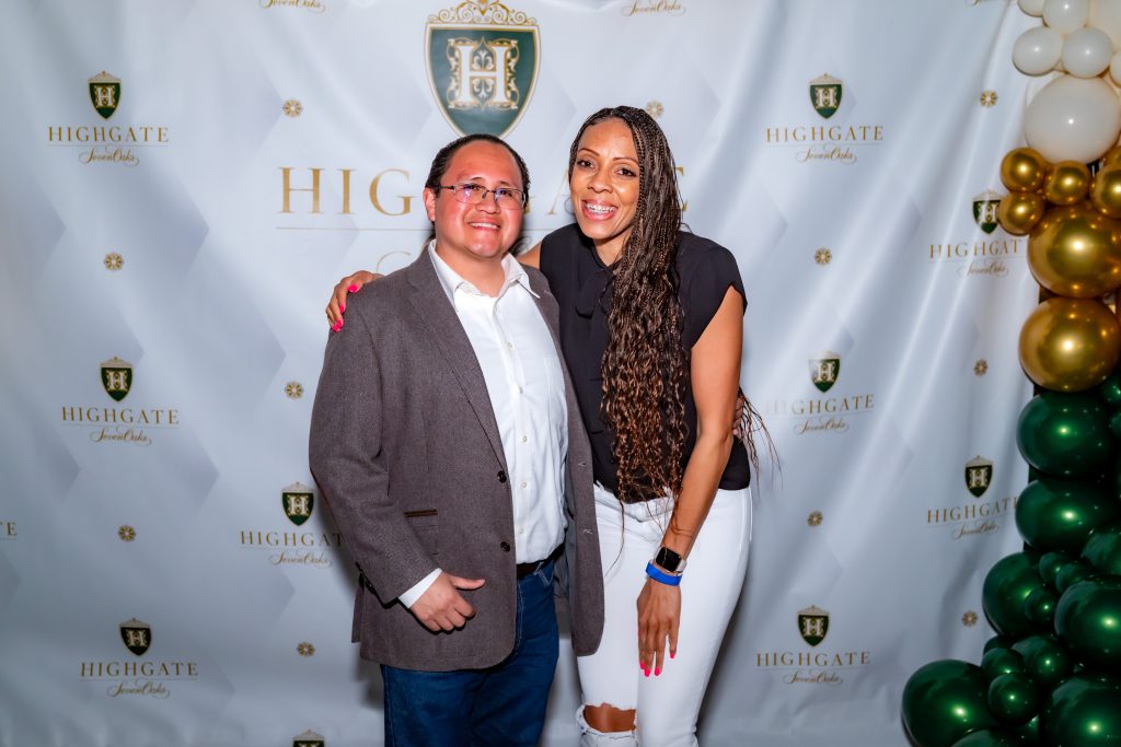 A man in a blazer and jeans stands next to a woman in a black shirt and ripped white jeans, both smiling in front of a "Highgate Jewels" step-and-repeat banner decorated with balloons.