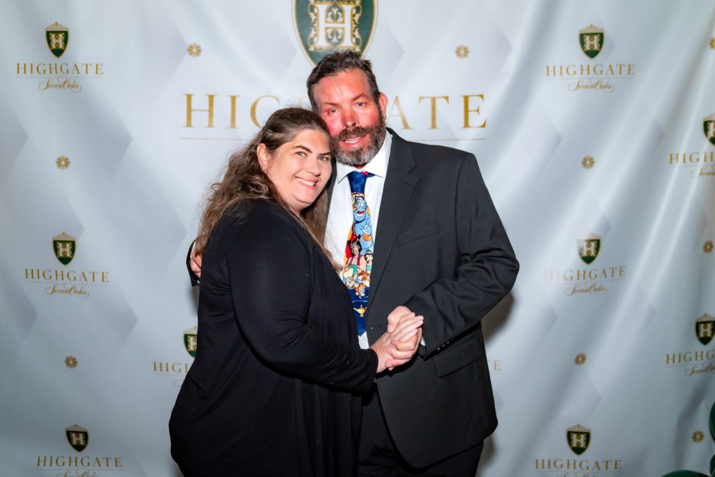A woman and a man in formal attire smile and pose together in front of a step-and-repeat backdrop with the "Highgate" logo. The man wears a suit and colorful tie; the woman is dressed in black.