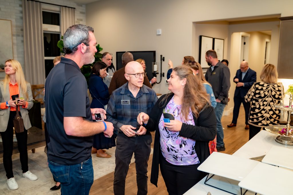 A group of people mingle and chat at an indoor social event. Some hold drinks and small boxes, while others talk in small groups. The atmosphere is warm and lively, with modern decor in the background.