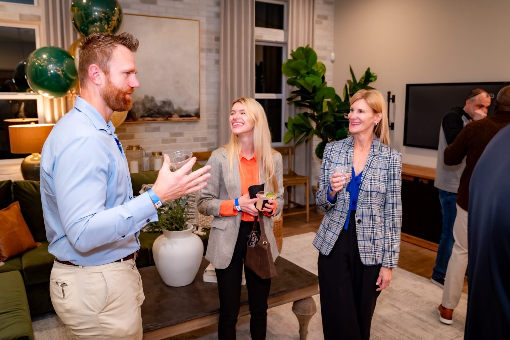 Three people stand in a modern living room, smiling and chatting at a social gathering. Two women hold drinks and a phone, while a man gestures as he talks. Balloons and plants decorate the cozy, well-lit space.