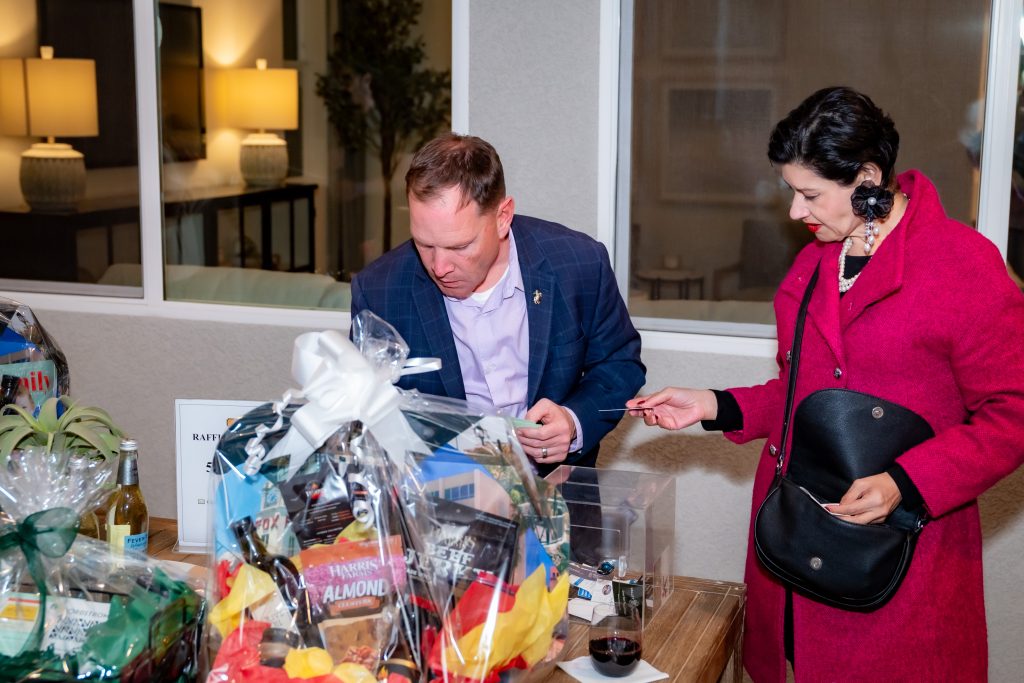 A man in a suit and a woman in a red coat examine a raffle table with gift baskets. The woman holds a ticket and wears a headset, while the man looks inside a box on the table. Indoor setting with soft lighting.